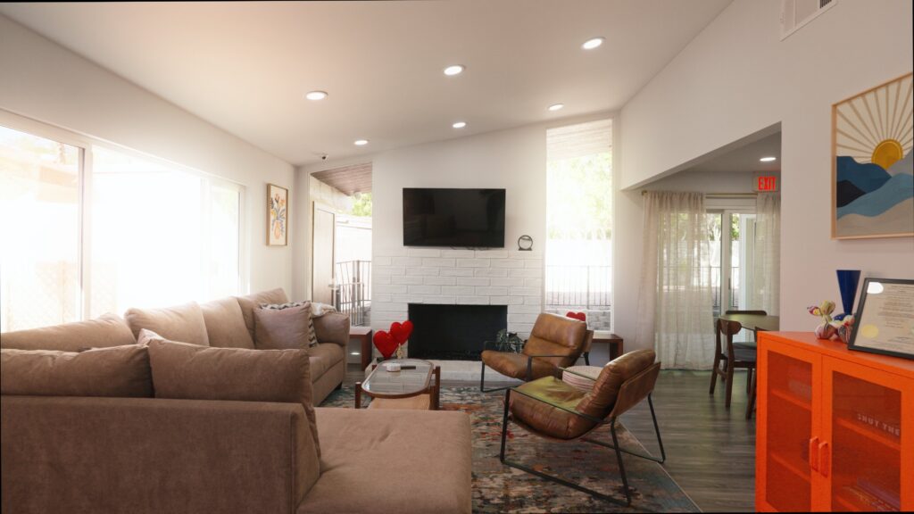 Living room with a large beige sectional sofa, two brown leather chairs, a glass coffee table, and a white brick fireplace with a wall-mounted TV above it. The room features a colorful area rug, large windows with sheer curtains, and an orange cabinet with decorative items and framed artwork on top. The walls are white, and recessed ceiling lights illuminate the space.