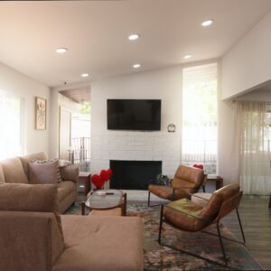 Living room with a large beige sectional sofa, two brown leather chairs, a glass coffee table, and a white brick fireplace with a wall-mounted TV above it. The room features a colorful area rug, large windows with sheer curtains, and an orange cabinet with decorative items and framed artwork on top. The walls are white, and recessed ceiling lights illuminate the space.