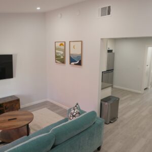 Living room with a teal sofa and a round wooden coffee table on a light rug. A flat-screen TV is mounted on the wall above a wooden media console decorated with a blue vase, a framed photo, and a banana. Two framed landscape paintings hang on the adjacent wall. The space opens into a dining area with a small round wooden table and four chairs, and a kitchen area with a stainless steel trash can and refrigerator. Light wood flooring and white walls throughout.