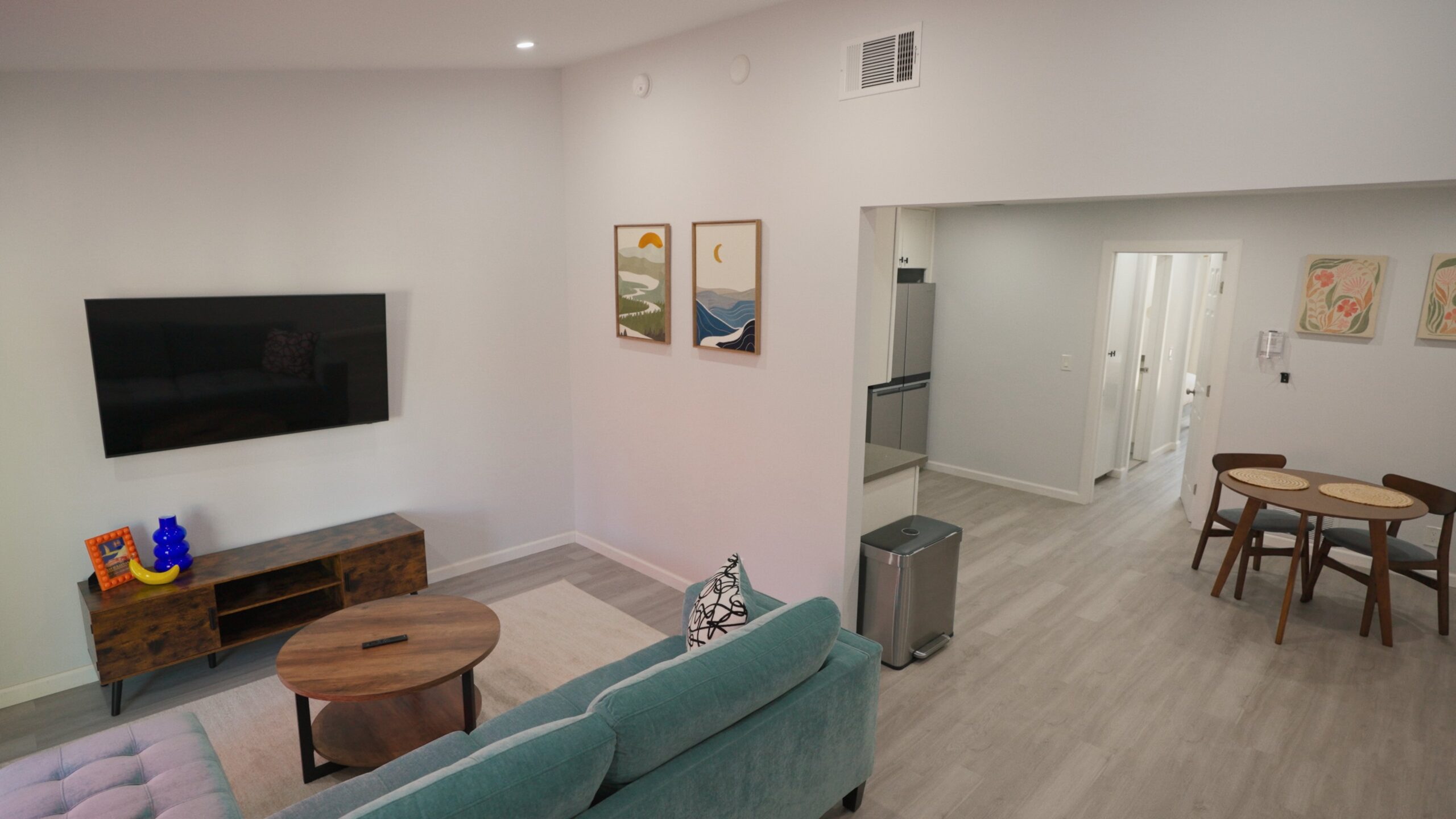 Living room with a teal sofa and a round wooden coffee table on a light rug. A flat-screen TV is mounted on the wall above a wooden media console decorated with a blue vase, a framed photo, and a banana. Two framed landscape paintings hang on the adjacent wall. The space opens into a dining area with a small round wooden table and four chairs, and a kitchen area with a stainless steel trash can and refrigerator. Light wood flooring and white walls throughout.