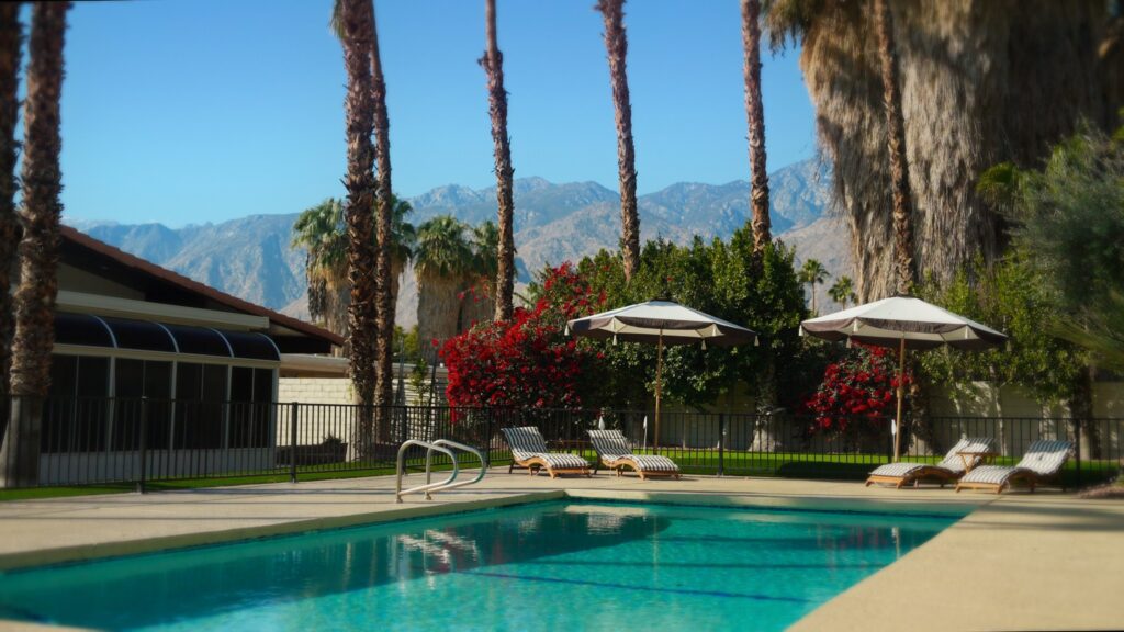 Outdoor swimming pool with clear blue water, surrounded by a concrete deck. Four striped lounge chairs are arranged near two large beige umbrellas. Tall palm trees and vibrant red flowering bushes line the background, with mountains visible in the distance under a clear blue sky. A black metal fence encloses the pool area.