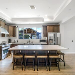 Modern kitchen with dark wood cabinets, stainless steel appliances including a large stove and refrigerator, a white countertop island with five cushioned bar stools, and a window above the sink overlooking an outdoor wooden structure. The floor is light wood, and there is a sliding glass door leading to an outdoor seating area.