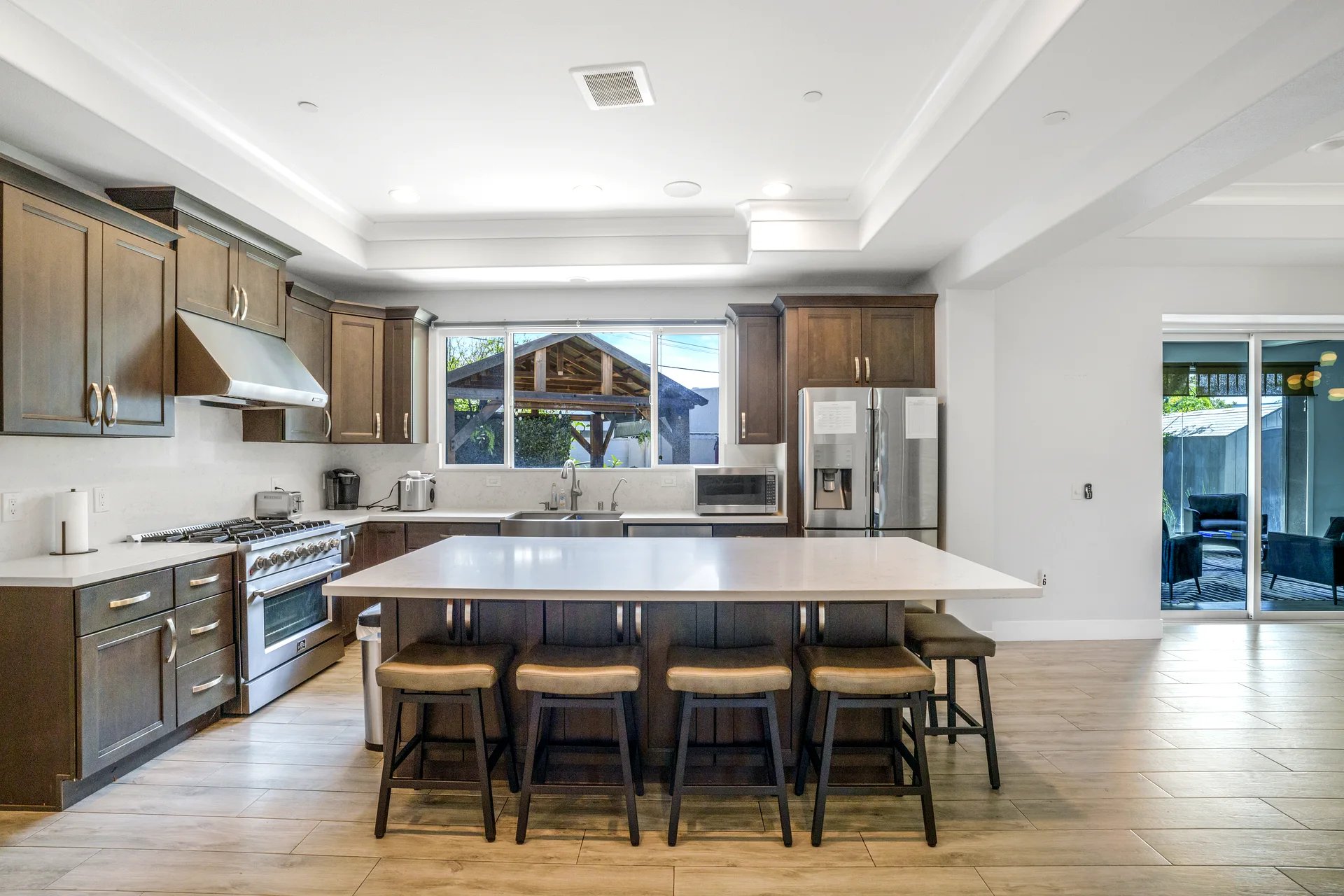 Modern kitchen with dark wood cabinets, stainless steel appliances including a large stove and refrigerator, a white countertop island with five cushioned bar stools, and a window above the sink overlooking an outdoor wooden structure. The floor is light wood, and there is a sliding glass door leading to an outdoor seating area.