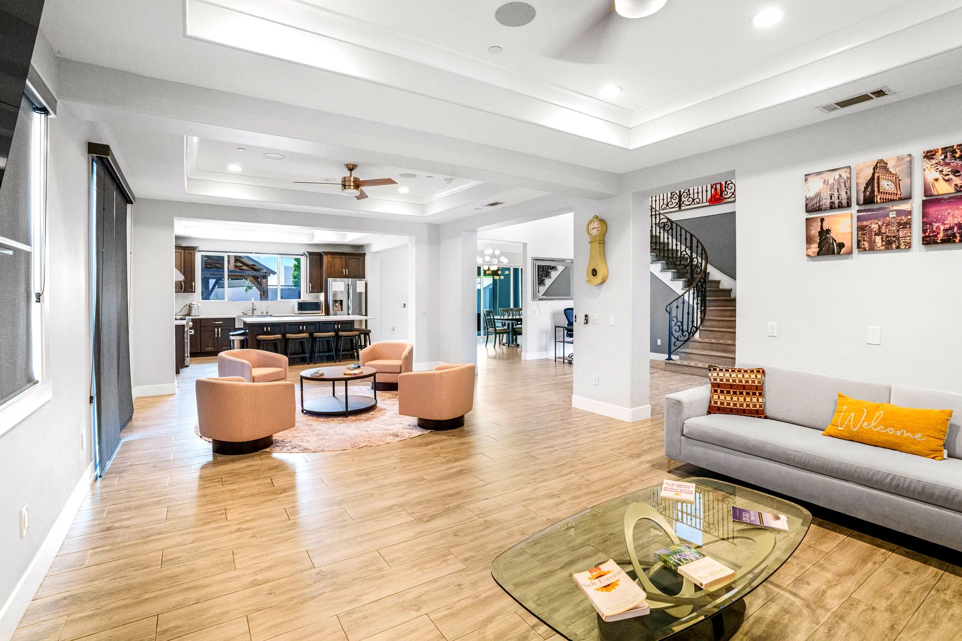Open-concept living area with light wood flooring, featuring a seating arrangement of four peach-colored armchairs around a round coffee table on a circular rug. To the right, a gray sofa with patterned and yellow pillows sits next to a glass coffee table holding several books. The kitchen in the background has dark wood cabinets, a large island with bar stools, and stainless steel appliances. A spiral staircase with ornate black railing is visible near the wall adorned with framed cityscape photos. The ceiling has recessed lighting and ceiling fans.