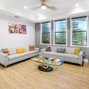 Modern living room with two light gray sofas, each adorned with colorful throw pillows including yellow and patterned designs. A glass coffee table with a unique curved base sits in the center on light wood flooring. Three large windows with dark roller shades allow natural light and show greenery outside. The walls are painted white with a gray accent wall behind the windows. A ceiling fan with a light fixture is mounted on the white tray ceiling. A set of six framed cityscape photos is displayed on one wall, and a textured green pouf is placed near the corner.