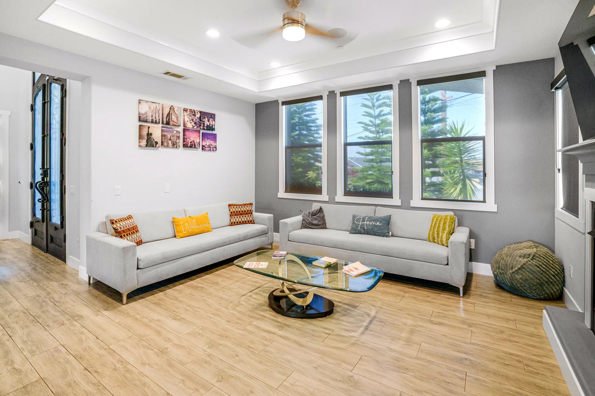 Modern living room with two light gray sofas, each adorned with colorful throw pillows including yellow and patterned designs. A glass coffee table with a unique curved base sits in the center on light wood flooring. Three large windows with dark roller shades allow natural light and show greenery outside. The walls are painted white with a gray accent wall behind the windows. A ceiling fan with a light fixture is mounted on the white tray ceiling. A set of six framed cityscape photos is displayed on one wall, and a textured green pouf is placed near the corner.