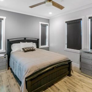 Bedroom with a dark wooden bed frame and a beige comforter. The bed has white and striped pillows, including a decorative pillow with the word "LOVE" on it. The walls are painted in two tones: gray behind the bed and light gray on the adjacent walls. There are four windows with black roller shades, white trim, and two mirrored dressers on either side of the bed. The floor is light wood, and a ceiling fan with wooden blades and a light fixture is mounted on the white ceiling.