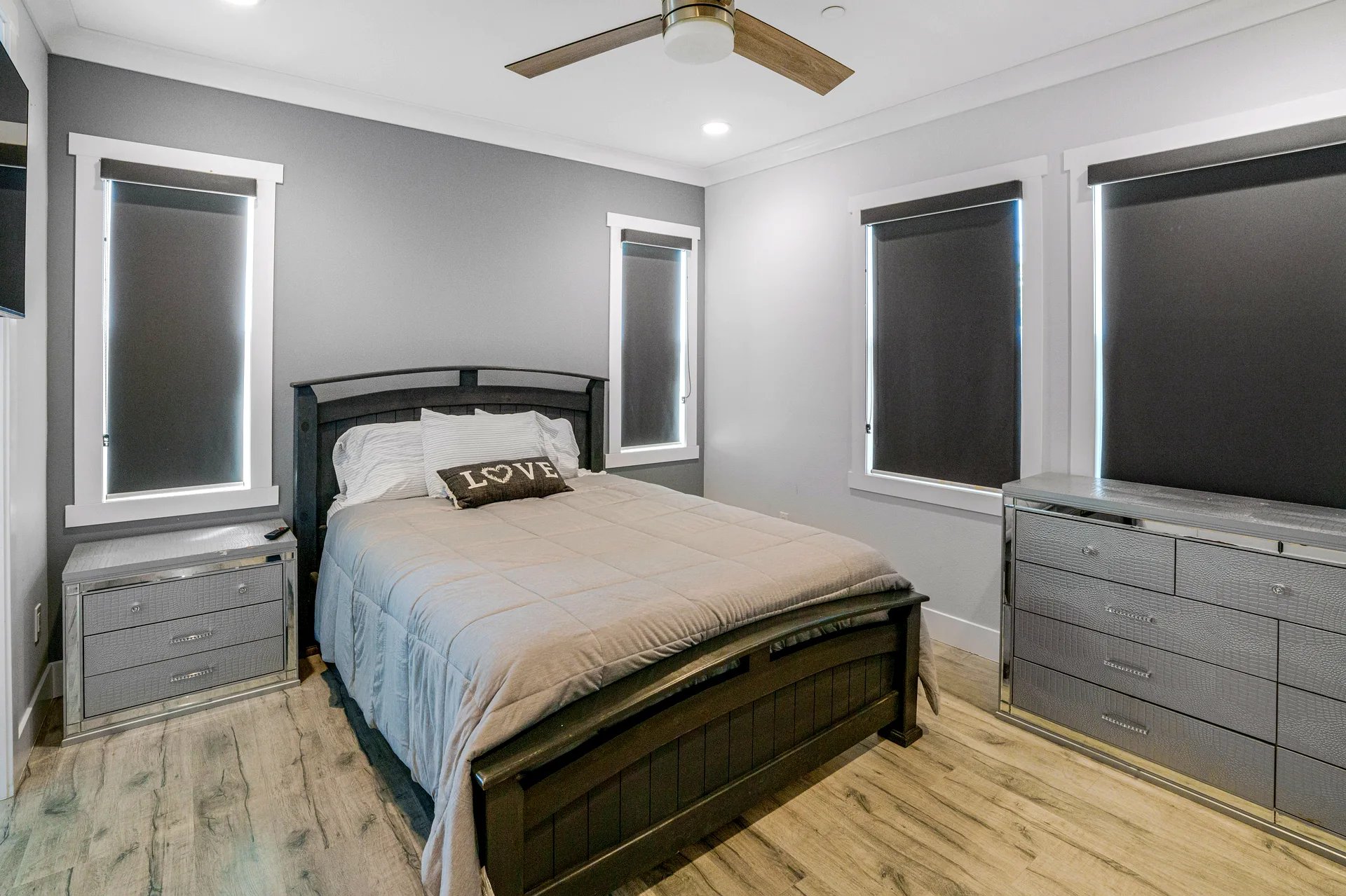 Bedroom with a dark wooden bed frame and a beige comforter. The bed has white and striped pillows, including a decorative pillow with the word "LOVE" on it. The walls are painted in two tones: gray behind the bed and light gray on the adjacent walls. There are four windows with black roller shades, white trim, and two mirrored dressers on either side of the bed. The floor is light wood, and a ceiling fan with wooden blades and a light fixture is mounted on the white ceiling.