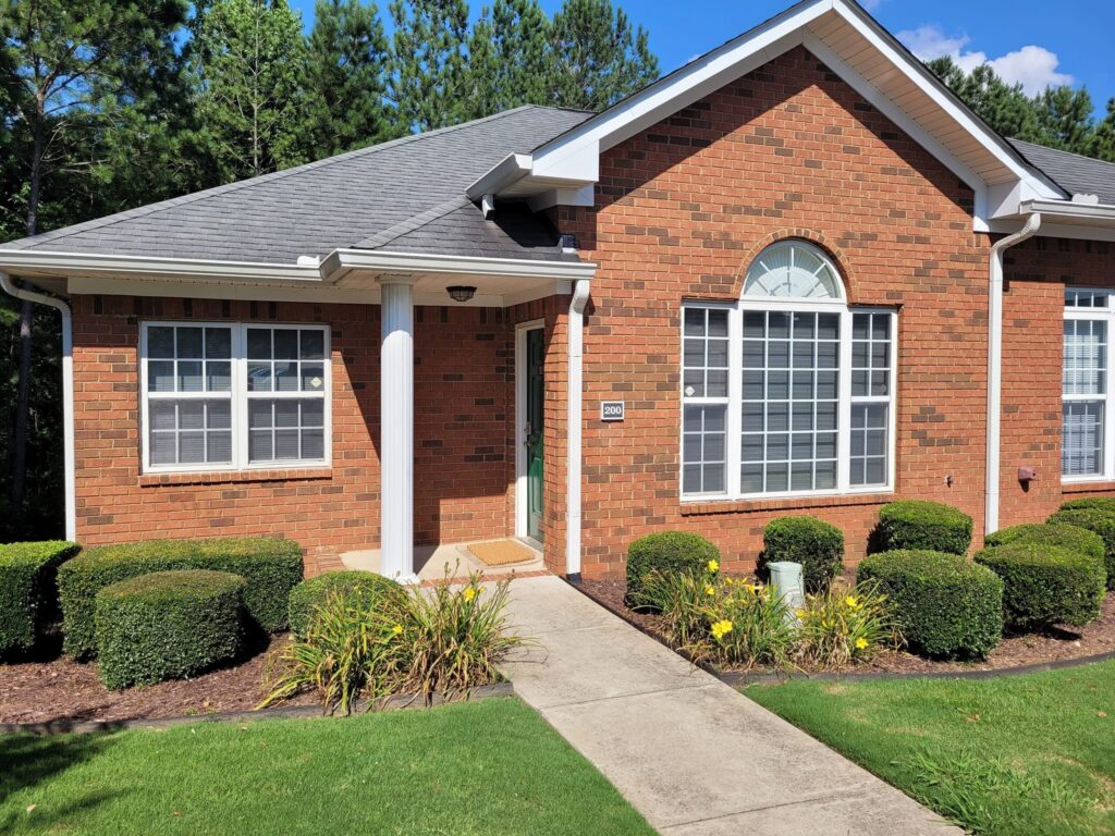 Brick house with a gray shingled roof, white trim, and multiple white-framed windows, including a large arched window. The entrance features a green door under a small covered porch supported by a white column. Well-maintained landscaping with neatly trimmed bushes and yellow flowers lines the concrete walkway leading to the door. Tall pine trees are visible in the background under a clear blue sky.
