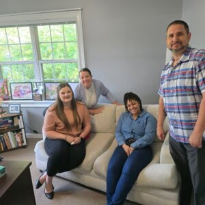 Four people are in a cozy office or living room setting. Two women are seated on a light beige couch, one wearing a peach top and black pants, the other in a blue denim shirt and dark pants. Another woman, dressed in a light gray cardigan and white top, leans on the back of the couch. A man in a blue, white, and red plaid short-sleeve shirt and dark pants stands to the right. Behind them is a window with a view of green trees, and a wooden bookshelf with books and framed pictures. The room has light gray walls and a carpeted floor.