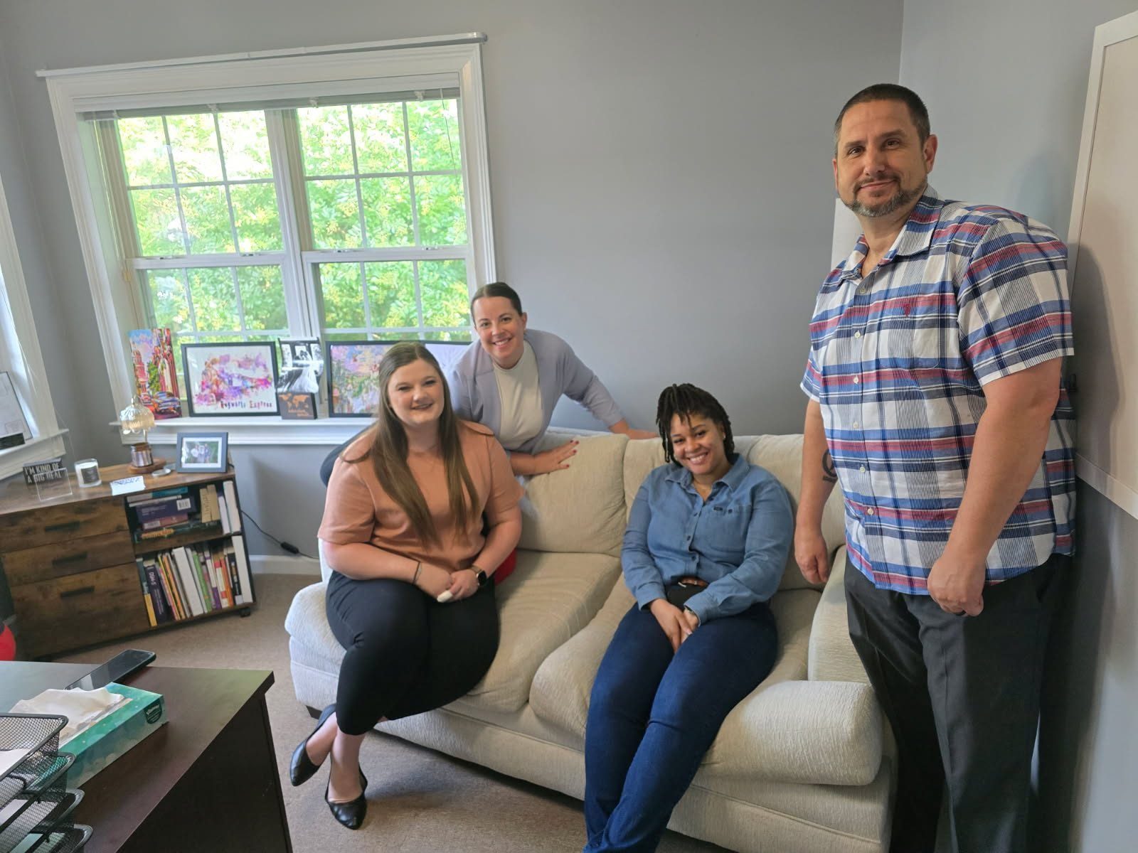 Four people are in a cozy office or living room setting. Two women are seated on a light beige couch, one wearing a peach top and black pants, the other in a blue denim shirt and dark pants. Another woman, dressed in a light gray cardigan and white top, leans on the back of the couch. A man in a blue, white, and red plaid short-sleeve shirt and dark pants stands to the right. Behind them is a window with a view of green trees, and a wooden bookshelf with books and framed pictures. The room has light gray walls and a carpeted floor.