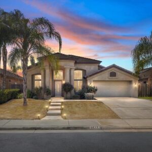 Single-story house with a beige exterior, a two-car garage, and a front entrance flanked by two large columns. The front yard features a concrete walkway with small lights, dry grass, and several palm trees. The sky is colorful with a sunset glow.