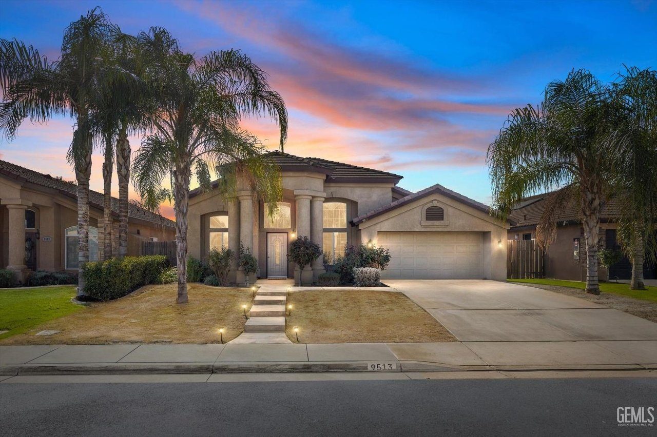 Single-story house with a beige exterior, a two-car garage, and a front entrance flanked by two large columns. The front yard features a concrete walkway with small lights, dry grass, and several palm trees. The sky is colorful with a sunset glow.