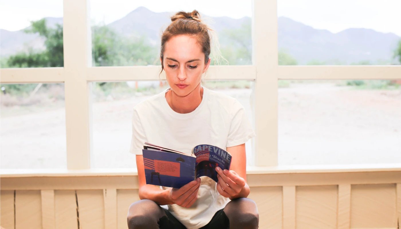 A woman with her hair tied up is sitting and reading a booklet titled "GRAPEVINE." She is wearing a plain white t-shirt and dark pants. The background shows large windows with an outdoor view of trees and mountains.
