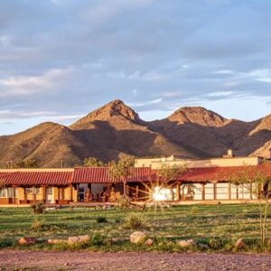A long, single-story building with a red-tiled roof and beige walls sits in front of a range of rugged, sunlit mountains. The foreground features a grassy area with scattered small plants and rocks, and a wooden teepee-like structure is visible on the left side. The sky above is partly cloudy with soft light illuminating the scene.