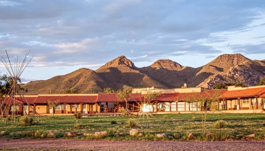 A long, single-story building with a red tile roof and beige walls sits in front of a range of rugged, sunlit mountains. The building has a covered porch area with wooden beams and several windows and doors. In the foreground, there is a grassy area with scattered small plants and rocks, and a wooden teepee-like structure made of tall poles on the left side. The sky above is partly cloudy with soft light illuminating the scene.