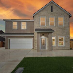 Two-story house with a stone facade on the right side and a beige stucco finish on the left. The house features a double garage with a white door, six windows on the front, and a central entrance with a small covered porch. The driveway and walkway are concrete, and there is a green lawn in front. The sky is a dramatic orange and yellow sunset.
