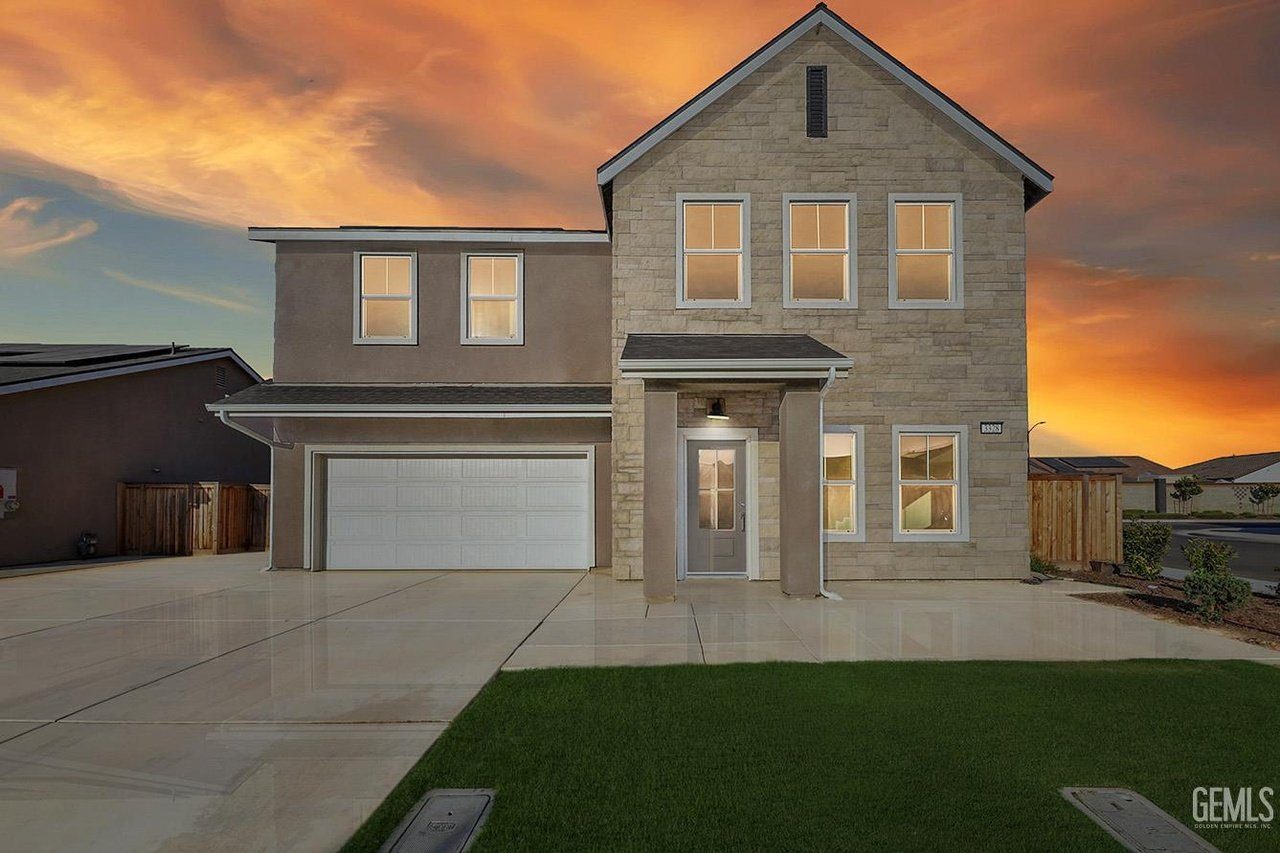 Two-story house with a stone facade on the right side and a beige stucco finish on the left. The house features a double garage with a white door, six windows on the front, and a central entrance with a small covered porch. The driveway and walkway are concrete, and there is a green lawn in front. The sky is a dramatic orange and yellow sunset.