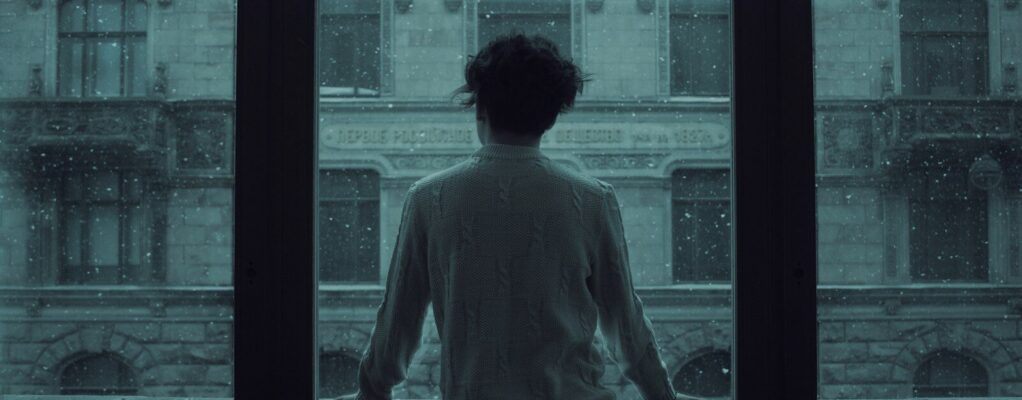 A person with short, curly hair wearing a textured knit sweater is standing indoors, facing away, and looking out through large windows at a historic stone building with arched windows. Snow is falling outside, creating a serene and contemplative atmosphere.