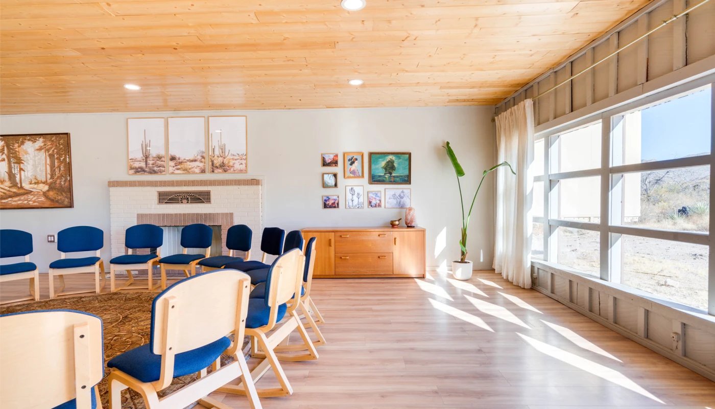 Room with a wooden ceiling and light wood flooring, featuring a circle of wooden chairs with blue cushions around a brown patterned rug. A white brick fireplace is centered on the far wall, flanked by framed artwork. To the right, a wooden sideboard holds decorative items, and a tall potted plant stands near large windows with sheer white curtains, allowing sunlight to stream in.