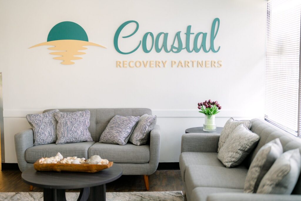 Waiting room with gray sofas, patterned pillows, coffee tables, flowers, and a wall sign reading “Coastal Recovery Partners.”
