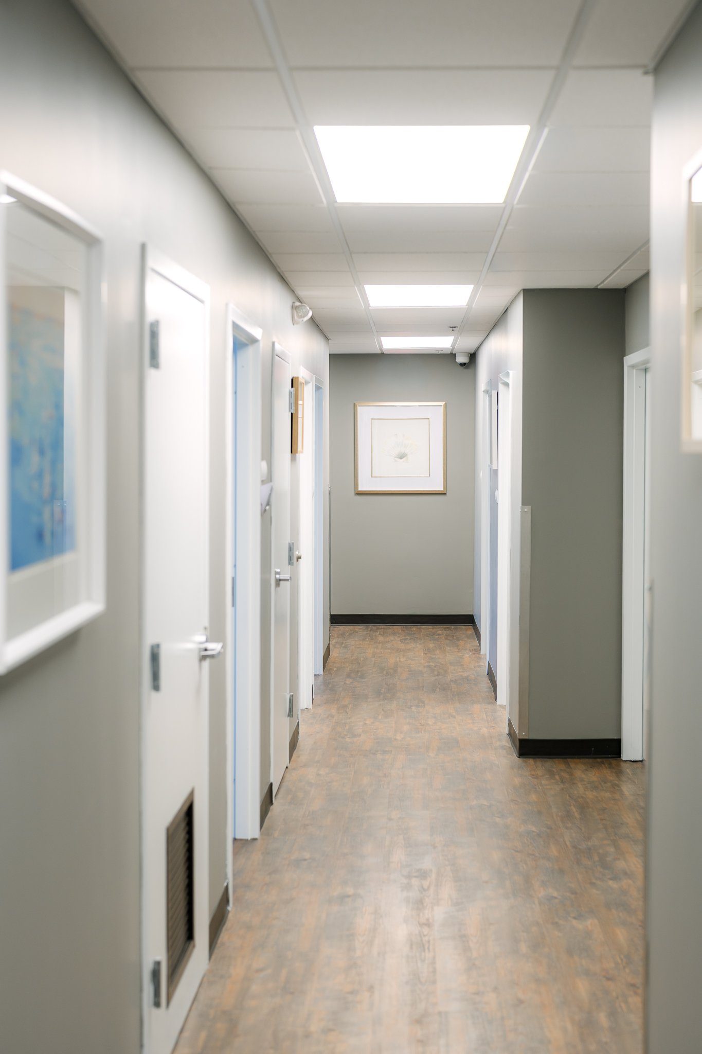 Empty office hallway with white doors, framed wall art, and fluorescent ceiling lights