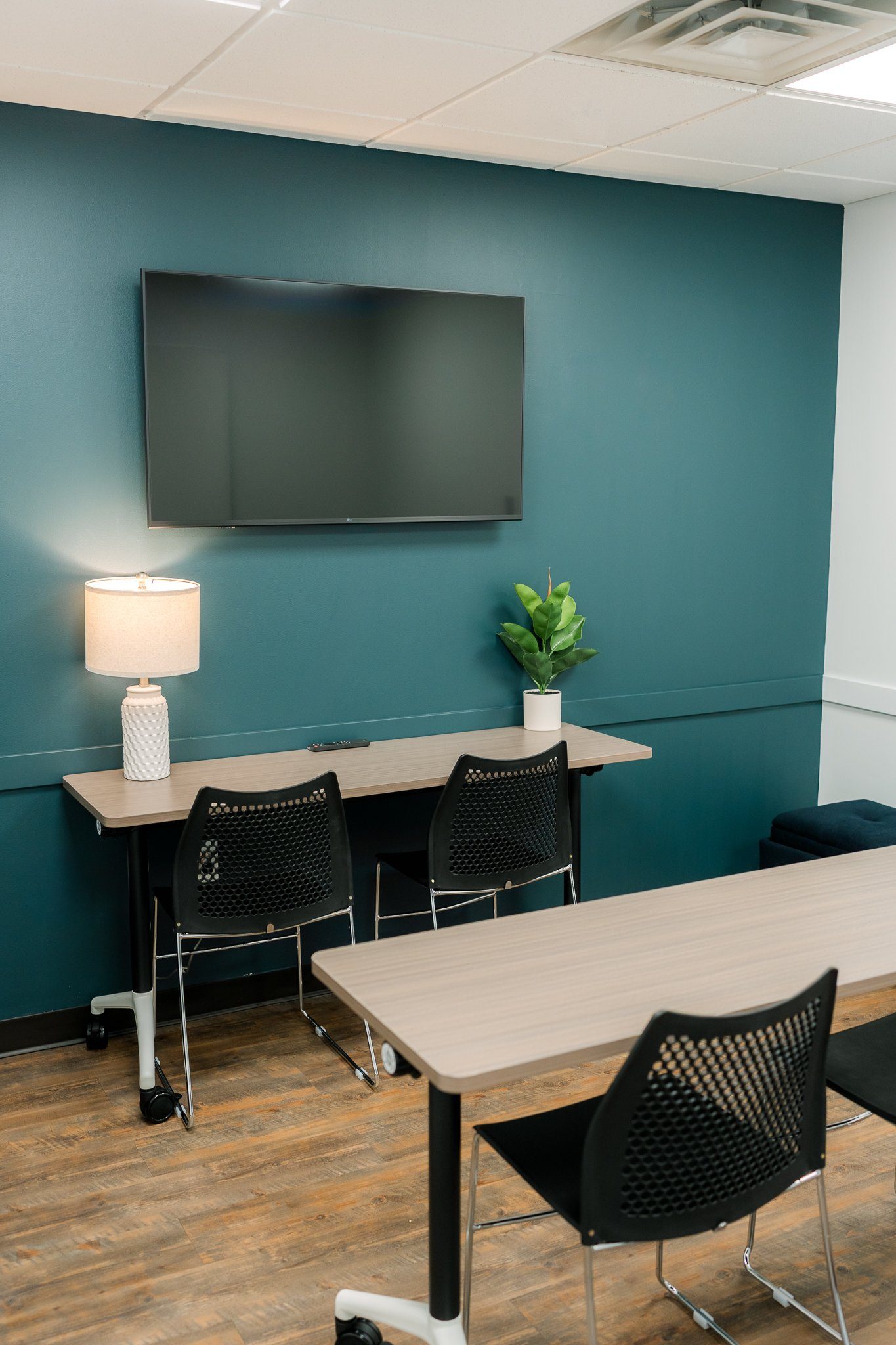 Office meeting room with wall-mounted TV, lamp, potted plant, tables, and black chairs against a teal wall.
