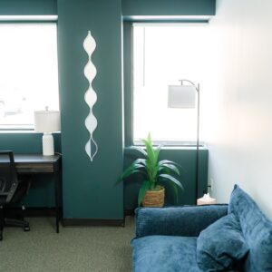Small office lounge with teal walls, desk and lamp, potted plant, floor lamp, and blue sofa near windows.