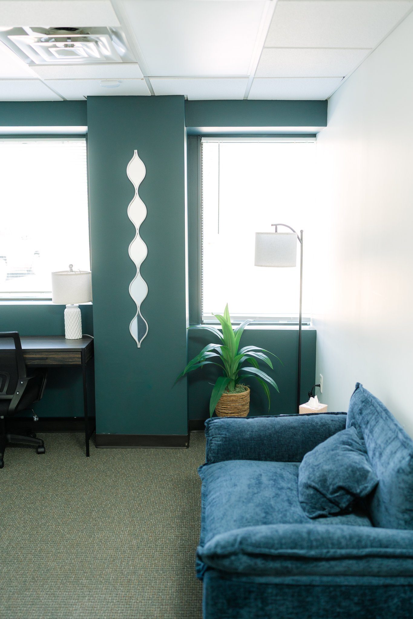 Small office lounge with teal walls, desk and lamp, potted plant, floor lamp, and blue sofa near windows.
