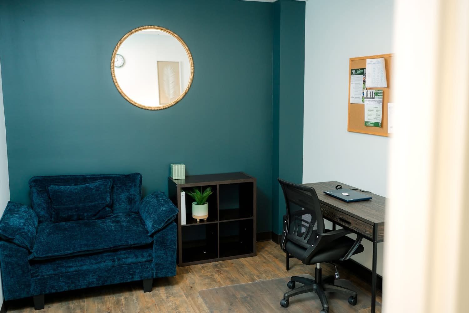 Small office with teal walls, blue sofa, desk chair, desk, cubby shelf, round mirror, and bulletin board.