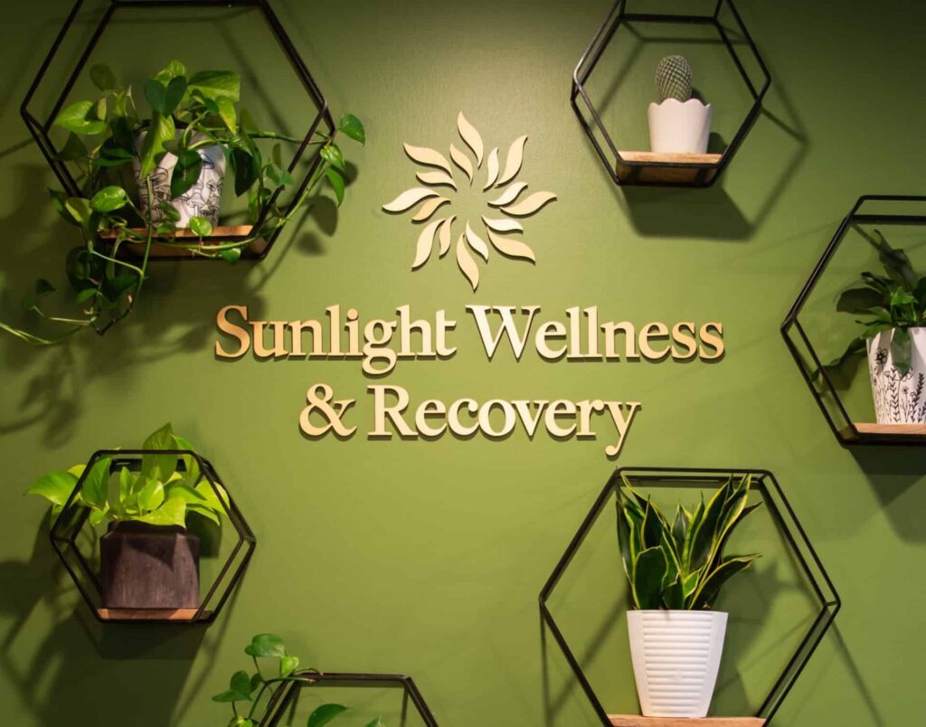 Green wall with hexagon shelves holding potted plants and the sign “Sunlight Wellness & Recovery” in the center
