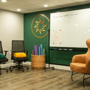 Office meeting room with whiteboard, rolling chairs, yoga mats, and a tan armchair on a green wall.