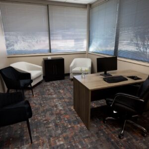 Small office with desk, computer, black chair, two armchairs, and windows with blinds