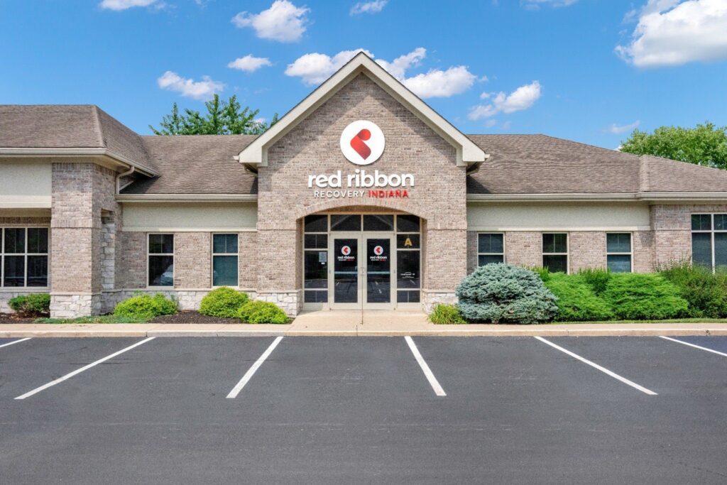 Brick office building with “red ribbon RECOVERY INDIANA” sign, glass entrance, and empty parking spaces under a blue sky
