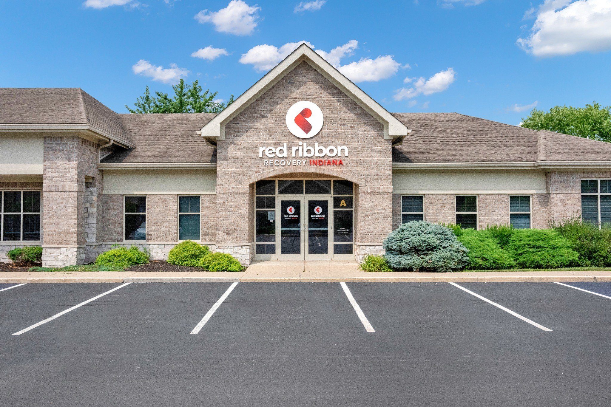 Brick office building with “red ribbon RECOVERY INDIANA” sign, glass entrance, and empty parking spaces under a blue sky