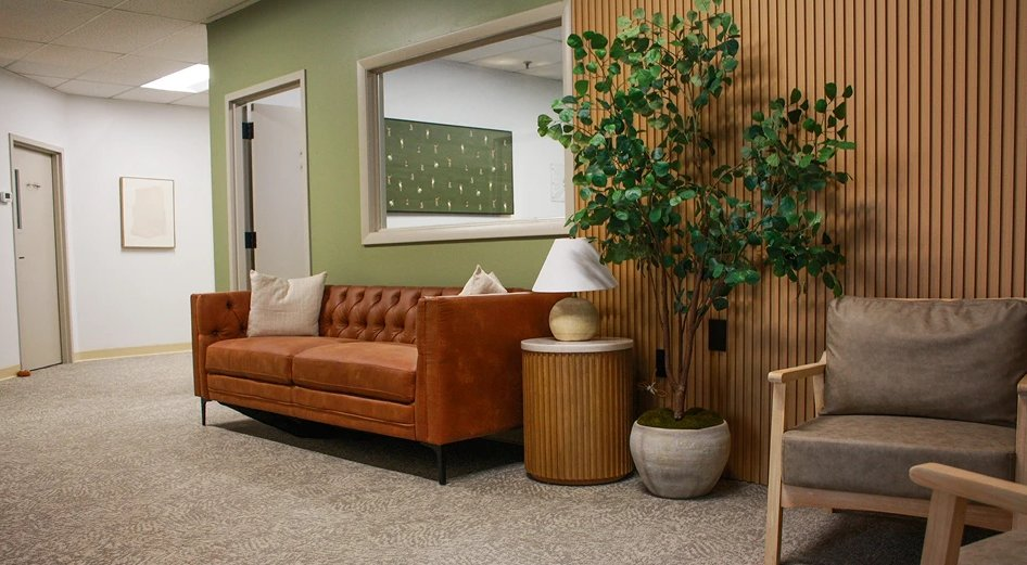 Modern waiting area with a brown sofa, gray chair, potted tree, side table, and wood slat wall.