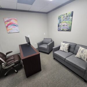 Small office with desk, leather chair, gray armchair and sofa, and two colorful wall paintings.