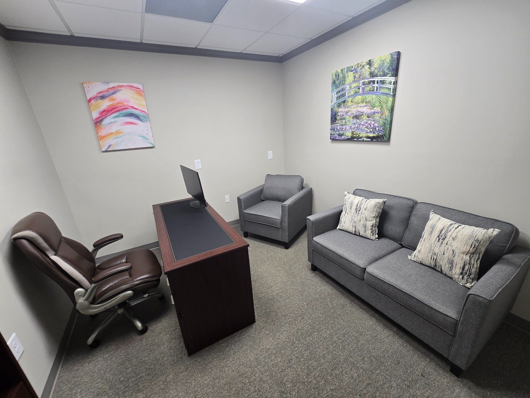 Small office with desk, leather chair, gray armchair and sofa, and two colorful wall paintings.