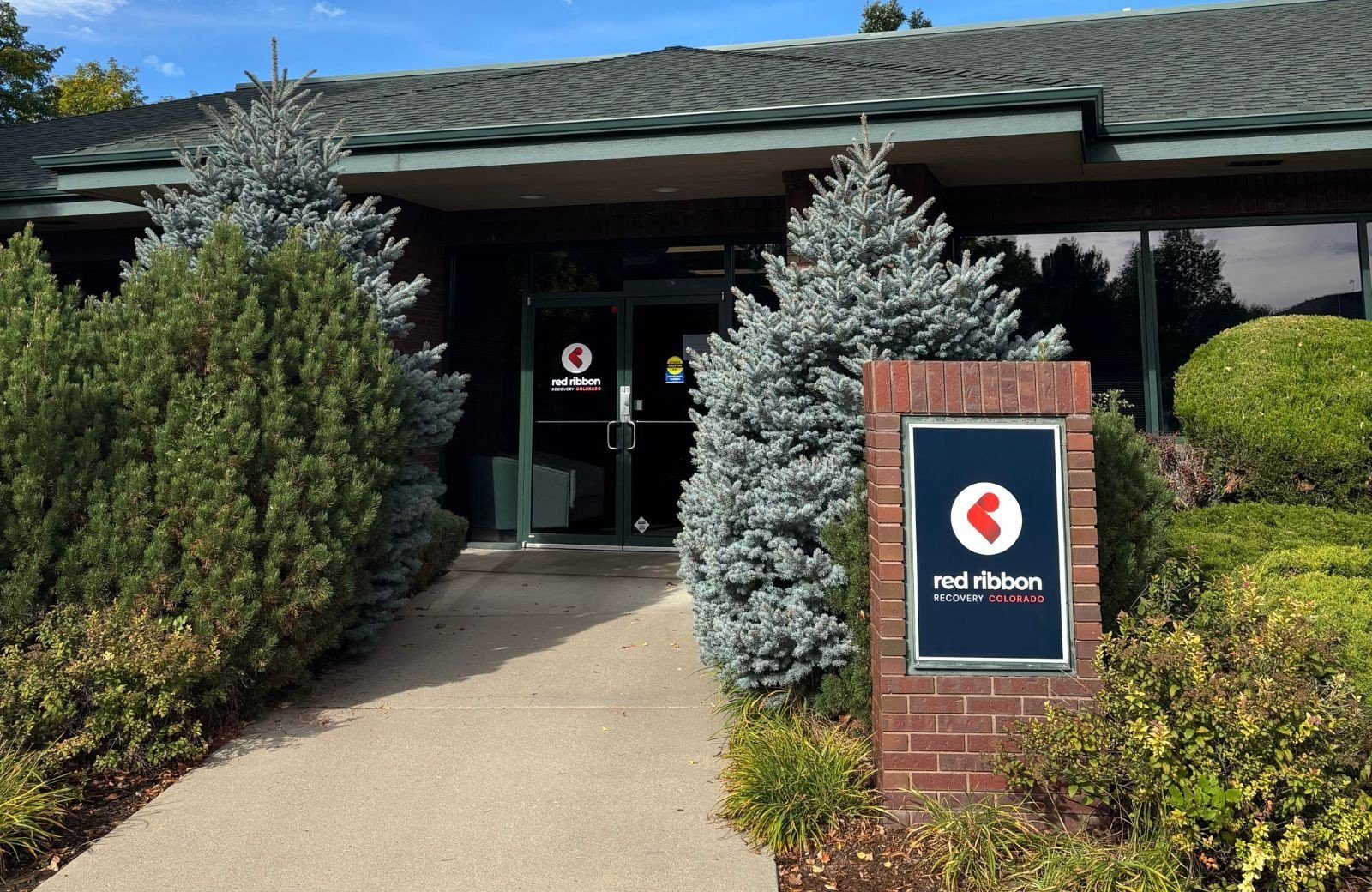 Entrance to a building with a brick sign reading “red ribbon recovery colorado” and glass doors behind shrubs and trees